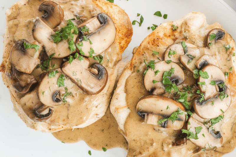 A Texas Roadhouse Smothered Chicken presentation. On a white plate, there are two pieces of chicken topped with mushrooms and a creamy sauce, garnished with chopped parsley.